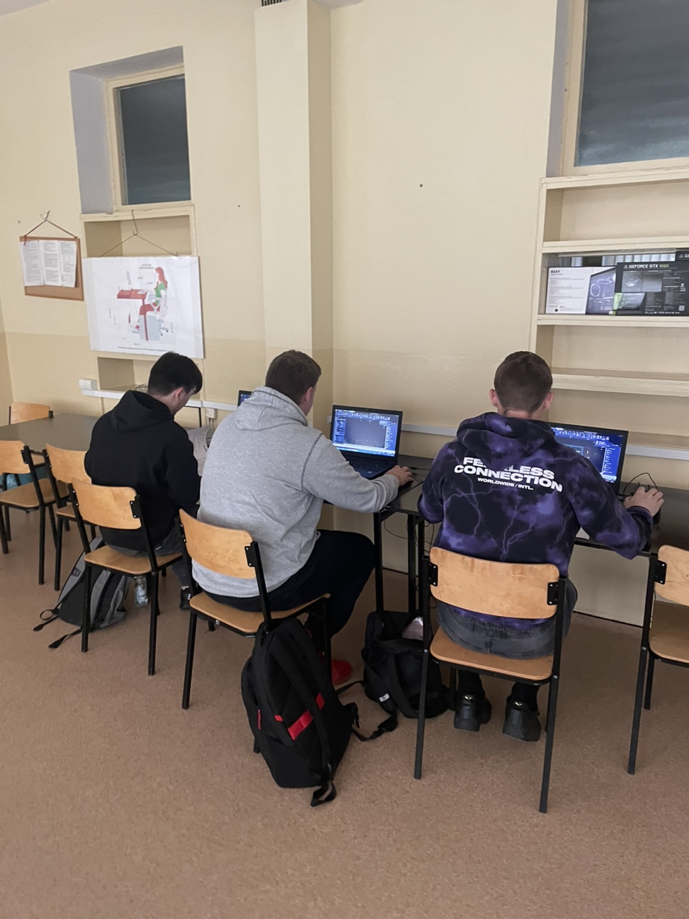 Trzy osoby siedzące przy komputerach w szkolnej sali komputerowej.